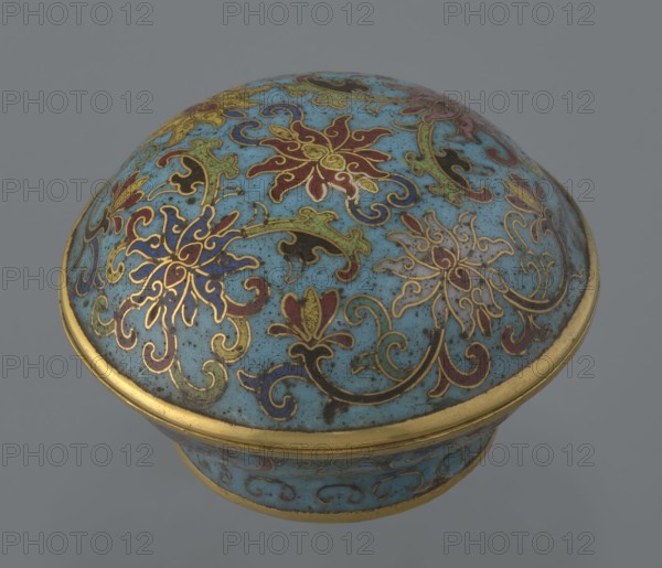 Box with a lid, cloisonné enamel, Qianlong period Qing dynasty, 18th century, China. Cylindrical spheroidal box on a separate ring foot with a slightly convex lid, decorated overall with multi-coloured lotus tendrils on a turquoise ground; foot with a band of curling silkworm-larvae motif. Rounded edge of lid, body rim, lower edge of foot, interior and bottom gilded. Bottom bears an engraved signature made in the Qianlong era and a workshop/guild mark yue. Pair of two similar items that differ slightly in shape, height, and ornamentation. Container, height 4 cm, diameter 6.7 cm., A rounded decorative object with a domed lid and a short cylindrical base, featuring an overall light blue background with ornate floral and scrollwork patterns outlined in gold; the motifs include stylized flowers and leaves in colors such as red, dark blue, green, black, and pale yellow, with gold borders along the rim and base, and the surface shows small speckling and subtle signs of wear.