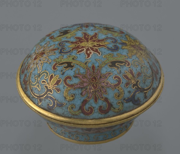 Box with a lid. Cylindrical, spheroidal shape on a separate ring foot, with a slightly convex lid. Entire surface decorated in cloisonné enamel with multi-coloured lotus tendrils on a turquoise ground. Foot bears a band with curling silkworm-larvae motif. Rounded edge of the lid, the storage section, lower edge of the foot, interior and bottom are gilded. Bottom engraved with an inscription: made in the Qianlong era, and a workshop/guild mark yue. Qing dynasty, Qianlong period 18th century, China. Height 4 cm; diameter 6.7 cm., A rounded decorative container lid with a domed top featuring intricate floral and scroll patterns in blue, gold, red, green, purple, and black, set against a speckled light blue background; the edge of the lid and a band near the base have gold trim, and the overall object shows fine metalwork lines and ornamental flourishes.