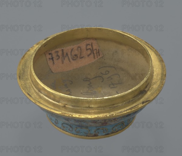 Box with a lid, Chinese, Qing dynasty, Qianlong period 1736–1795, cloisonné enamel, cylindrical spheroidal shape on a separate ring foot, slightly convex lid. Entire surface decorated with multi-coloured lotus tendril on a turquoise ground; foot bears a band with curling silkworm-larva motif. Rounded edge of lid, rim of box, lower edge of foot, interior and bottom gilded. Bottom engraved with inscription made in the Qianlong era and workshop/guild mark yue. Pair with a similar item; two pieces differ slightly in shape, height, and ornamentation. 18th century, China. Container; height 4 cm, diameter 6.7 cm., A shallow circular metal vessel with a wide rim, showing a worn golden surface and a few small dark spots, sits against a gray background; the interior base has a muted beige tone with some faint smudging and a small irregular patch of brownish paper, and the exterior band near the base displays a decorative turquoise area with swirling gold patterns and touches of darker blue and g...