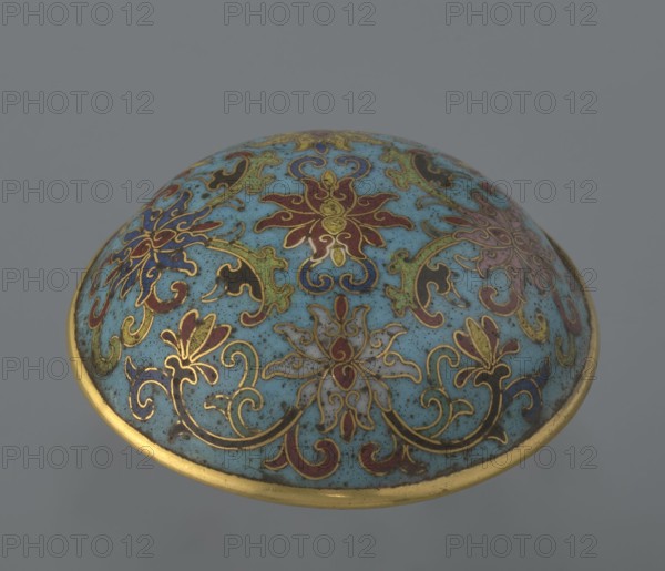 Cylindrical cloisonné enamel box with lid, spheroidal shape on a separate ring foot, slightly convex lid, decorated overall with multi-coloured lotus tendril ornament on a turquoise background. Foot bears a band with a curling silkworm-larva decorative motif. Rounded edge of lid, storage section, lower edge of foot, interior and bottom are gilded. Bottom bears an engraved signature made in the Qianlong era and a workshop/guild mark yue. Pair with a similar item that differs slightly in shape, height, and ornamentation. Qing dynasty, Qianlong period 18th century, China. Height 4 cm; diameter 6.7 cm. Materials: cloisonné enamel., A rounded ornamental object with a domed surface decorated in a light blue background and ornate floral and scroll patterns in gold outlines filled with colors including red, dark blue, green, purple, black, and muted beige, with a gold rim around the edge.
