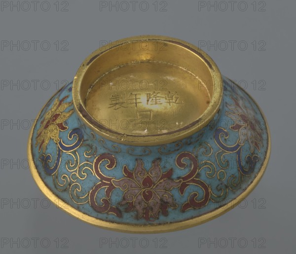 Cylindrical spheroidal box with lid, cloisonné enamel on metal, 18th century, China, Qianlong period 1736–1795. Cylindrical box on a separate ring foot with a slightly convex lid, fully decorated with multi-coloured lotus tendrils on a turquoise ground. Foot band with curling silkworm-larvae motif. Rounded edge of lid, lower edge of foot, interior and bottom gilt. Bottom engraved: made in the Qianlong era, with workshop/guild mark yue. Paired with a similar box slightly different shape, height, and ornamentation. Height 4 cm; diameter 6.7 cm., A shallow, rounded metal vessel with a raised central rim, showing a gold interior and gold edging, sits against a gray background; the exterior surface is covered in a turquoise enamel with ornate floral and scrolling patterns outlined in gold and filled with shades of red and purple, and there are a few worn areas where the gold and turquoise show slight discoloration.