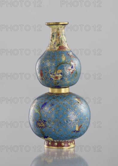Double gourd-shaped vase, cloisonné enamel, decorated with flowers, butterflies, and insects on a turquoise ground with a grid of circles, yellow neck. Qing dynasty 19th century, China. Vessel, height 14 cm, diameter 7 cm. Vase twisted from two parts., A double-gourd shaped vase with a smooth surface in shades of blue and gold, featuring a pattern of interlocking gold lines and floral motifs in colors including red, purple, green, and white; the neck area transitions to a yellowish tone with additional decorative swirls in similar colors, and the base ring shows a band in a reddish color with gold accents; the object sits against a plain light gray background and is reflected faintly below.
