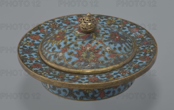 Tin with a lid, Chinese, Qing dynasty, Qianlong period, cloisonné enamel. Cylindrical tin with a wide plate-like collar extending beyond the vessel walls, slightly rising at the rim. Domed lid with a handle in the shape of a decorative knob with openwork relief: a dragon in clouds, its head directed toward a pearl atop the handle. Exterior box and collar decorated with a multicolored red, blue, green, yellow, white small-flowered ornament — branches with rosettes, buds, and leaves — on a turquoise background. Lid decorated with a lotus tendril composed of four geometrically arranged flowers with surrounding branches; a multi-petal rosette in the center under the metal handle, also on turquoise. Bottom filled with a rich multicolored composition of elements reminiscent of Buddhist symbols — a vajra and Dharma wheel forming the inner rosette, with two-colored red and blue ribbons between the symbols. Interior of box and lid without enamel decoration; all interior elements gilded. Possibl...