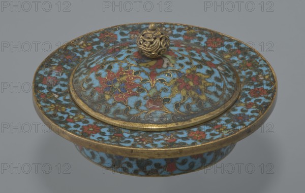 Tin with a lid, Chinese, Qing dynasty Qianlong period, cloisonné enamel on tin, 18th century. Cylindrical tin with a wide plate-like collar extending beyond the vessel walls and slightly rising at the rim. Domed lid with a handle in the form of a decorative openwork knob: a dragon in clouds with its head directed toward a pearl atop the handle. Exterior of the vessel box and collar decorated with a multicolored red, blue, green, yellow, white small-flowered ornament — branches with rosettes, buds, and leaves — on a turquoise background. Lid decorated with a lotus tendril composed of four geometrically arranged flowers and surrounding branches; a multi-petal rosette at the center under the metal handle, on turquoise background. Bottom filled with a rich multicolored composition of elements reminiscent of Buddhist symbols — a vajra and Dharma wheel forming the inner central rosette, with two-colored red and blue ribbons between the symbols. Interior of box and lid unenameled and gilded; ...