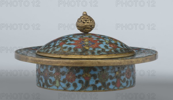Tin with a lid, Chinese, Qing dynasty, Qianlong period, cloisonné enamel, 18th century. Cylindrical tin with a wide plate-like collar extending beyond the vessel walls, slightly rising at the rim. Domed lid with a handle in the form of an openwork dragon in clouds, head directed toward a pearl atop the handle. Exterior of box and collar decorated with multicolored red, blue, green, yellow, white small-flowered ornament—branches with rosettes, buds, and leaves—on a turquoise background. Lid decorated with a lotus tendril of four geometrically arranged flowers and surrounding branches; a multi-petal rosette at the center beneath the metal handle, all on turquoise. Base filled with a rich multicolored composition of elements reminiscent of Buddhist symbols—a vajra and Dharma wheel forming the central rosette—with two-colored red and blue ribbons between the symbols. Interior of box and lid unenameled; all interior elements gilded. Possibly used in religious ritual incense box. Container; ...