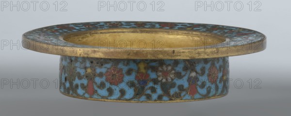 Tin with a lid;, Chinese, Qing dynasty, Qianlong period, 18th century. Cloisonné enamel on tin; cylindrical container with a wide plate-like collar extending beyond the vessel walls and slightly rising at the rim; domed lid with a decorative openwork knob depicting a dragon in clouds facing a pearl. Exterior box and collar decorated with multicolored red, blue, green, yellow, white small-flowered ornament — branches with rosettes, buds, and leaves — on a turquoise background. Lid decorated with a lotus tendril composed of four geometrically arranged flowers and surrounding branches; a multi-petal rosette centered beneath the metal handle, also on turquoise. Bottom filled with a rich multicolored composition of elements reminiscent of Buddhist symbols a vajra and Dharma wheel forming the central rosette with two-colored red and blue ribbons between the symbols. Interior of box and lid gilt without enamel. Possibly used as a ritual incense box. Height 7.7 cm; diameter 15.1 cm., A shallow...