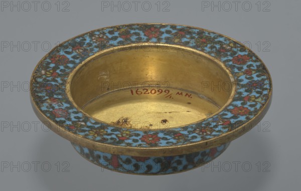 Tin with a lid, 18th century, China. Cloisonné enamel on tin. Cylindrical tin with a wide plate-like collar extending beyond the vessel walls, slightly rising at the rim. Domed lid with a handle shaped as a decorative openwork relief: a dragon in clouds, head directed toward a pearl at the top. Exterior of the vessel box and collar decorated with a multicolored red, blue, green, yellow, white small-flowered ornament—branches with rosettes, buds, and leaves—on a turquoise background. Lid decorated with a lotus tendril of four geometrically arranged flowers and surrounding branches; a multi-petal rosette is centered under the metal handle, all on a turquoise ground. Bottom filled with a rich, multicolored composition evoking Buddhist symbols—a vajra and Dharma wheel forming the inner central rosette—with two-colored red and blue ribbons between the motifs. Interior of box and lid without enamel decoration; all interior elements gilded. Possible function: incense box. Height 7.7 cm; diame...
