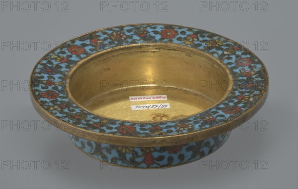Tin with a lid, Chinese art, Chinese culture, Qing 1644–1911, Qianlong period, cloisonné enamel; cylindrical tin with a wide plate-like collar extending beyond the vessel walls and slightly rising at the rim. Domed lid with a decorative knob handle in openwork relief: a dragon in clouds with its head directed toward a pearl at the top of the handle. Exterior box and collar decorated with a multicolored red, blue, green, yellow, white small-flowered ornament — branches with rosettes, buds, and leaves — on a turquoise background. Lid decorated with a lotus tendril composed of four geometrically arranged flowers and surrounding branches; center beneath the metal handle has a multi-petal rosette, also on turquoise. Bottom filled with a rich multicolored composition of elements reminiscent of Buddhist symbols — a vajra and Dharma wheel forming the inner central rosette, with two-colored red and blue ribbons between the Buddhist symbols. Interior of box and lid devoid of enamel decoration; a...