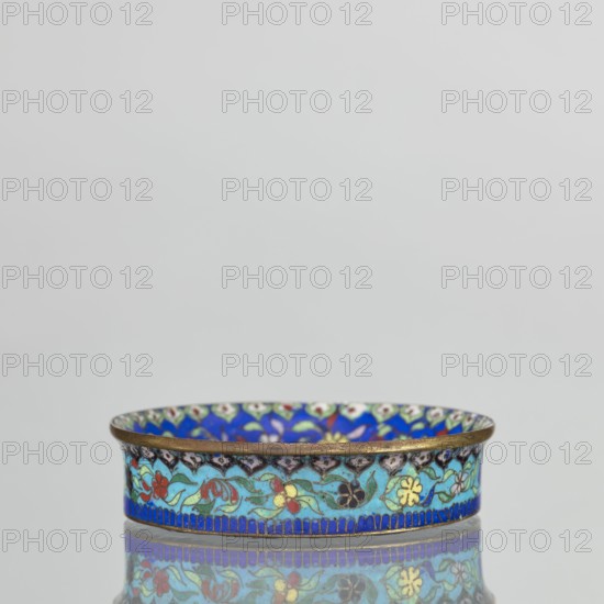 Bowl, gift, Chinese art, Chinese, Qing 1644–1911, decorative vessel, cloisonné enamel, flowers, butterflies; decoration: inside the bottom decorated with flowers on a light lilac background, collar decorated with flowers on a sapphire background; outside the collar decorated with flowers on a turquoise background, bottom covered with lilac enamel. 19th century, China, vessel; height 2.3 cm, diameter 9.5 cm., A shallow round dish with an outwardly curved rim, decorated with a repeating floral pattern in bright colors; the exterior band shows stylized flowers and leaves in shades of turquoise, blue, green, red, yellow, black, and white set against a blue border, with a thin metallic rim at the top; the interior surface visible at the rim carries a similar multicolored floral motif with dominant blue and green tones; the dish sits on a reflective surface that mirrors its colors and pattern, and the background is a plain light gray.