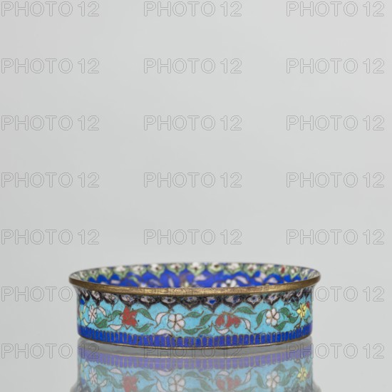 Bowl, 19th century, China. Cloisonné enamel decorative vessel, height 2.3 cm, diameter 9.4 cm. Round, flat-bottomed bowl with low side wall widening toward a curved, gilded outward rim. Mirror decorated on a light purple ground around a central black rosette with four butterflies in white, red, and black, and multicolored flowers including Buddha's fingers/Chinese lemon and surrounding leaves arranged in a circle. Inner wall has a floral tendril on a blue background with multicolored flowers similar to the mirror decoration; a lambrequin ornament at the edge resembles rui-head shapes. Outer wall in turquoise carries a decoration similar to the inner wall. Underside of the bottom coated with single-color purple enamel. Paired with another bowl., A shallow circular dish with a metallic rim decorated with a repeating floral enamel pattern in blues, turquoise, green, red, white, yellow and black, set against a neutral pale background and showing a reflected image of the dish on a glossy su...