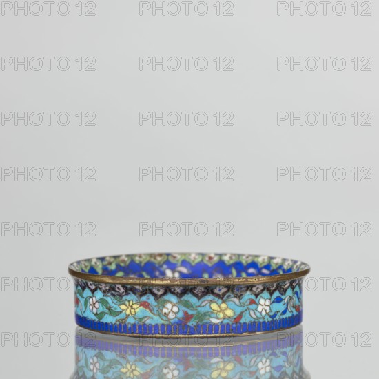 Bowl, 19th century, China. Cloisonné enamel decorative vessel, height 2.3 cm, diameter 9.4 cm. The round, flat-bottomed bowl with a low side wall widening toward the rim is decorated in the mirror with an ornamental composition on a light purple background arranged around a central black rosette. The composition includes four butterflies in white, red, and black, multicolored flowers including Buddha's fingers/Chinese lemon and surrounding leaves arranged in a circle. The inner wall is decorated with a floral tendril against a blue background composed of multicolored flowers similar to those in the mirror. A lambrequin ornament at the edge resembles the shape of rui heads. The rim is curved outward and gilded. The turquoise outer wall is filled with decoration similar to that on the inner wall. The underside of the bottom is covered with a single-color purple enamel. Pair with another bowl. Qing dynasty. Chinese., A shallow round metal dish with a decorative rim sits centered against a...