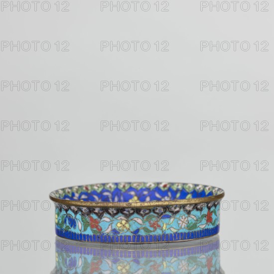 Bowl, gift; Chinese; Qing dynasty 1644–1911; cloisonné enamel decorative vessel, 19th century; height 2.3 cm, diameter 9.4 cm. The round, flat-bottomed bowl with a low side wall widening toward the rim is decorated in the mirror with an ornamental composition on a light purple background arranged around a central black rosette. The composition comprises four butterflies in white, red, and black, multicolored flowers including Buddha’s fingers/Chinese lemon and surrounding leaves arranged in a circle. The inner wall is decorated with a floral tendril on a blue background of multicolored flowers similar to those in the mirror. A lambrequin ornament is visible at the edge, reminiscent of the shape of rui heads. The rim curves outward and is gilded. The turquoise outer wall bears decoration similar to the inner wall. The underside of the bottom is covered with a single-color purple enamel. This piece forms a pair with another bowl., A shallow circular dish with a metallic rim decorated wit...