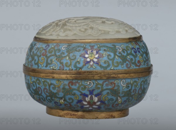 Box with a lid, Chinese, Qing dynasty, Qianlong period, cloisonné enamel on metal with a slightly convex bas-relief grayish jade plaque on the lid. Oval spheroidal box on a ring foot, divided halfway into container and lid. Jade plaque depicts a fenghuang phoenix or peacock with a sun among clouds; the sun is upper left surrounded by a silk ribbon. Motifs and styling reference emblems from mandarin squares: peacock indicating third civil rank and the sun referring to imperial authority. Plaque is a fine example of jade workmanship. Rounded sides of container and lid decorated with a multicolored lotus tendril band on a turquoise ground. Interior filled with uniform turquoise enamel. Metal rings reinforcing edges and plaque attachment are gilded. 18th century, China. Height 8.2 cm; width 11.3 cm; diameter 12.2 cm., A rounded decorative container with a domed carved lid sits against a plain pale gray background; the lid appears white and carved with raised swirling floral motifs and rest...