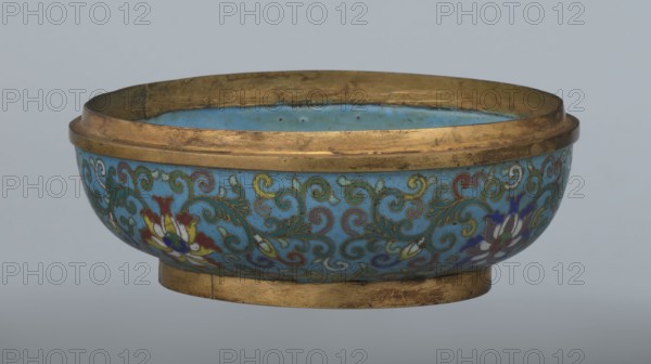 Box with a lid;, Chinese, Qing dynasty, Qianlong period, 18th century. Cloisonné enamel spheroidal box based on a ring foot, divided halfway into container and lid. Slightly convex bas-relief plaque of grayish jade set on the cover depicting a fenghuang phoenix or peacock with a sun among clouds; the sun is in the upper left surrounded by a silk ribbon. Motifs and styling reference emblems used on mandarin squares: a peacock signified third civil rank and the sun signified imperial authority. The jade plaque is a fine example of hard-stone workmanship. Rounded sides of both container and lid are decorated with a multicolored lotus tendril stripe on a turquoise ground. Interior filled with a uniform layer of turquoise enamel. Metal rings reinforcing the edges of the container, lid, and plaque attachment are gilded. Function: container. Dimensions: height 8.2 cm; width 11.3 cm; diameter 12.2 cm., A shallow round bowl with a wide rim and a low foot, showing a turquoise blue interior and e...