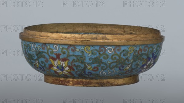 Box with a lid, cloisonné enamel, 18th century, China. Oval, spheroidal box on a ring foot, divided halfway into container and lid. Slightly convex bas-relief plaque of grayish jade set on top of the cover depicting a fenghuang phoenix or peacock with a sun among clouds; the sun is in the upper left corner and is surrounded by a silk ribbon. Motifs and styling refer to emblems appearing on mandarin squares: the peacock denoted third civil rank and the sun referred to imperial authority. The jade plaque is a fine example of hard-stone workmanship. Rounded sides of both container and lid are decorated with a multi-colored lotus tendril stripe on a turquoise ground. Interior filled with a uniform layer of turquoise enamel. Metal rings reinforcing the edges of the container, lid, and plaque attachment are gilded. Height 8.2 cm; width 11.3 cm; diameter 12.2 cm., A low, shallow bowl with a rounded body and a short foot, featuring a band of metallic gold around the rim and foot; the exterior ...