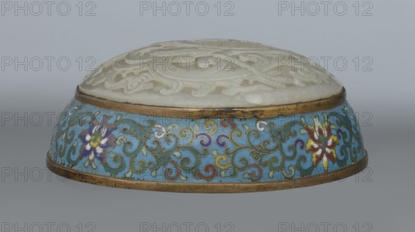 Box with a lid; cloisonné enamel on metal with a slightly convex grayish jade bas-relief plaque on the cover depicting a fenghuang phoenix or peacock with a sun among clouds; sun in upper left surrounded by a silk ribbon; motifs and styling reference emblems appearing on mandarin squares peacock indicating third civil rank; sun referring to imperial authority. Rounded sides of container and lid decorated with a multicolored lotus tendril stripe on a turquoise background. Interior filled with a uniform layer of turquoise enamel. Gilded metal rings reinforce edges of container, lid, and plaque attachment. Oval, spheroidal box based on a ring foot, divided halfway into container and lid. 18th century, Qing dynasty China. Height 8.2 cm; width 11.3 cm; diameter 12.2 cm., container function., A low round decorative object with a domed carved pale stone top showing swirling carved patterns, set into a thin metallic rim, sitting on a wider circular base decorated with intricate scrolling ename...