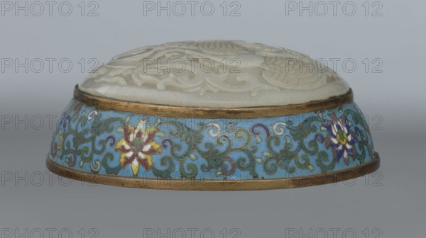 Box with a lid; cloisonné enamel on metal with a slightly convex bas-relief grayish jade plaque on top of the cover. Oval, spheroidal box based on a ring foot, divided halfway into container and lid. Plaque depicts a fenghuang phoenix or peacock with a sun among clouds; the sun is surrounded by a silk ribbon. Motifs and styling reference emblems appearing on mandarin squares: the peacock indicating third rank of a civilian official and the sun referring to imperial authority. The plaque is a finely worked example in jade. Rounded sides of both container and lid are decorated with a multicolored lotus tendril stripe on a turquoise background. Interior filled with a uniform layer of turquoise enamel. Metal rings reinforcing the edges of the container, lid, and plaque attachment are gilded. 18th century, China. Height 8.2 cm; width 11.3 cm; diameter 12.2 cm., A shallow round object with a domed carved top in off-white resting on a circular base decorated with a bright blue band, ornate gr...