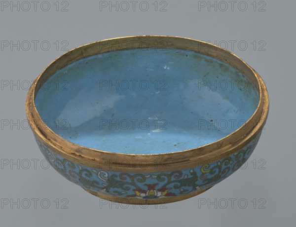 Box with a lid. Oval, spheroidal box on a ring foot, divided halfway into container and lid. Cloisonné enamel with gilded metal reinforcing rings; interior filled with uniform turquoise enamel. Slightly convex bas-relief grayish jade plaque added to top of cover, depicting a fenghuang phoenix or peacock with a sun among clouds; sun is upper left, surrounded by a silk ribbon. Motifs and styling reference emblems from mandarin squares: peacock denoting third civilian rank, sun referring to imperial authority. Rounded sides of both container and lid decorated with multi-colored lotus tendril stripes on a turquoise ground. Exceptionally fine jade workmanship noted; early precursor cloisonné technique used semi-precious stones set in metal cells. 18th century, China. Height 8.2 cm; width 11.3 cm; diameter 12.2 cm. Creator, : Paderewski, Ignacy Jan 1860–1941., A shallow round bowl with a smooth glossy interior in shades of blue and turquoise, bordered by a worn metallic rim in gold tones; th...