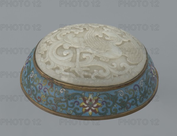 Box with a lid., Chinese, Qing dynasty, Qianlong period, 18th century. Cloisonné enamel on metal with a slightly convex bas-relief grayish jade plaque on the lid. Oval, spheroidal box based on a ring foot, divided halfway into container and lid. Lid plaque depicts a fenghuang phoenix or peacock with a sun among clouds; the sun is in the upper left surrounded by a silk ribbon. Motifs and styling reference mandarin square emblems peacock denoting third civilian rank; sun referring to imperial authority. Rounded sides of both container and lid decorated with multi-colored lotus tendril stripes on a turquoise ground. Interior filled with a uniform layer of turquoise enamel. Metal rings reinforcing the edges and the plaque attachment are gilded. Height 8.2 cm; width 11.3 cm; diameter 12.2 cm., A circular decorative object with a carved white top showing swirling raised patterns and bird-like shapes set into a shallow rounded base with a band of gold trim; the base features a turquoise backg...