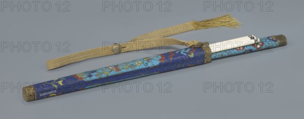 Travel cutlery set in case; Chinese, Qing dynasty 1644–1911, 19th century. Includes: a Knife with cloisonné enamel handle and steel blade. b–c Ivory chopsticks decorated at the top with an inlaid black lacquer ornament forming a border. d File decorated similarly to the chopsticks. e Case length 28 cm made of cloisonné enamel with bronze fittings engraved at top and bottom; interior lined with wood. Chopsticks and file are held by a band decorated with a border; a narrow cotton-silk strap with fringes is attached. Case decorated with floral reserves and plant tendrils in the background. Motifs include plant designs and a swastika iconography. Function: travel cutlery set., A long cylindrical case with decorative patterns lies diagonally across a gray surface, showing a deep blue base with swirls and floral motifs in lighter blue, red, yellow, green, and gold tones; the case has metallic end caps in an aged brass color and a matching metal band near the open end where a group of slender...