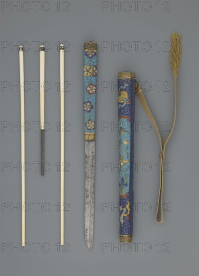 Travel cutlery set in case; Chinese, Qing dynasty 1644–1911, 19th century. Consists of: a Knife with cloisonné enamel handle and steel blade. b–c Ivory chopsticks, each decorated at the top with an inlaid black lacquer border ornament. d File decorated similarly to the chopsticks. e Case made of cloisonné enamel with bronze fittings engraved at top and bottom; interior lined with wood. Chopsticks and file held by a band decorated with a matching border; a narrow cotton-silk strap with fringes is attached. Case decorated with flowers in two reserves and plant tendrils in the background. Materials: cloisonné enamel, ivory, steel, bronze, lacquer. Function: travel cutlery. Length: 28 cm case. Iconography: plant motifs, swastika., A group of long slender objects arranged vertically on a gray background: a few pale ivory rods with smooth surfaces and darkened tips, one of which has a textured darker section partway down; a decorated cylindrical sheath and a matching scabbard both showing in...