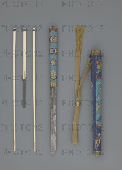 Travel cutlery set in case; Chinese, Qing dynasty 1644–1911, 19th century. Includes: a Knife with cloisonné enamel handle and steel blade. b–c Ivory chopsticks decorated with an inlaid black lacquer ornament at the top in the form of a border. d File decorated similarly to the chopsticks. e Case reaching 3/4 of the height of the cutlery; made of cloisonné enamel with bronze fittings engraved at top and bottom; interior lined with wood. Chopsticks and file are secured with a band decorated with a matching border; a narrow cotton-silk strap with fringes is attached. Case decorated with flowers in two reserves and plant tendrils in the background. Motifs include plants and a swastika. Cutlery; length 28 cm case., A group of thin, pale rods with dark tips arranged vertically on the left, a long narrow blade with a patinated metallic surface and a decorated cylindrical handle featuring floral patterns in blue, red, white, and gold near the center, a long woven strap in tan with a fringed en...