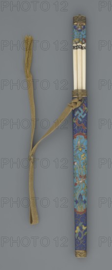 Travel cutlery set in case; 19th century, China, Qing dynasty. Knife with cloisonné enamel handle and steel blade; ivory chopsticks with inlaid black lacquer border ornament; file decorated similarly to the chopsticks. Case of cloisonné enamel with bronze fittings engraved at top and bottom, interior lined with wood; chopsticks and file held by a decorative band; narrow cotton-silk strap with fringes attached. Case decorated with flowers in two reserves and plant tendrils in the background. Plant motifs and swastika iconography present. Cutlery; length 28 cm case., A long cylindrical object with decorative enamel work in shades of blue, turquoise, red, green, gold, and a few touches of white, featuring floral and scrolling patterns and metal fittings at the ends; three smooth, pale rods protrude from the top, bound together with a band showing dark and light accents; an attached woven strap in tan with a knotted section and a tassel of loose fibers at its end lies alongside the cylindr...