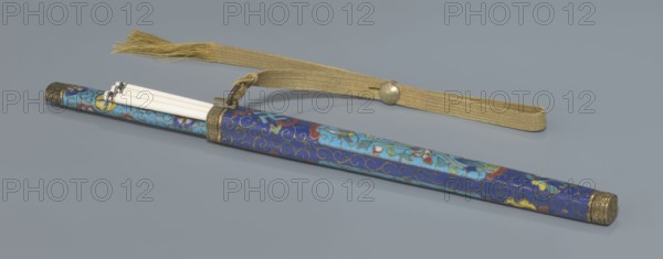 Travel cutlery set in case; Chinese, Qing dynasty 19th century. Knife with cloisonné enamel handle and steel blade. Ivory chopsticks inlaid with black lacquer border ornament. File decorated similarly to the chopsticks. Case of cloisonné enamel with bronze fittings engraved with ornamentation at top and bottom; interior lined with wood. Chopsticks and file held by a band with matching border; narrow cotton-silk strap with fringes attached. Case decorated with flowers in two reserves and plant tendrils in the background. Materials: cloisonné enamel, bronze, steel, ivory, lacquer, cotton-silk. Function: travel cutlery. Length: 28 cm case. Motifs: plant motifs and swastika iconography., A long cylindrical case with a decorative blue surface featuring swirling patterns and floral motifs in lighter blue, red, yellow, and green rests on a gray background, with metal end caps and a metal band near one end holding a few slender white sticks that extend partially out of the case; a gold-colored...
