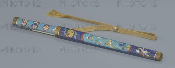 Travel cutlery set in case; 19th century, China Qing dynasty. Travel cutlery comprising: a knife with cloisonné enamel handle and steel blade; b–c ivory chopsticks decorated at the top with an inlaid black lacquer ornament in the form of a border; d file decorated similarly to the chopsticks; e cloisonné enamel case with bronze fittings engraved with ornamentation at top and bottom, interior lined with wood. Chopsticks and file are held by a band decorated with a matching border; a narrow cotton-silk strap with fringes is attached to the case. Case decorated with flowers in two reserves and plant tendrils in the background. Materials: cloisonné enamel, ivory, steel, bronze, lacquer; motifs: plant motifs, swastika; function: cutlery; length: 28 cm case., A long cylindrical object with decorative floral and scrollwork patterns lies diagonally across a flat surface alongside a woven strap with a tassel. The cylindrical piece shows areas of light blue and deep blue enamel with motifs in wh...