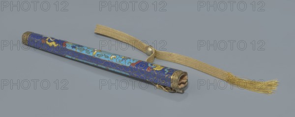 Travel cutlery set in case; 19th century, China, Qing dynasty. Consists of: a Knife with cloisonné enamel handle and steel blade. b–c Ivory chopsticks, each with an inlaid black lacquer border ornament at the top. d File decorated similarly to the chopsticks. e Case 28 cm length made of cloisonné enamel with bronze fittings engraved at top and bottom; interior lined with wood. Chopsticks and file held by a band with a matching border; narrow cotton-silk strap with fringes attached. Case decorated with flowers in two reserves and plant tendrils in the background. Plant motifs and swastika iconography present. Function: travel cutlery., A long cylindrical object lying diagonally on a smooth gray surface with a detached woven strap beside it; the cylinder is decorated in a patterned blue with lighter blue panels and accents of gold and red, with swirling gold lines and small curled motifs, and metal end caps that have a worn brass or bronze appearance; the woven strap is a golden beige co...
