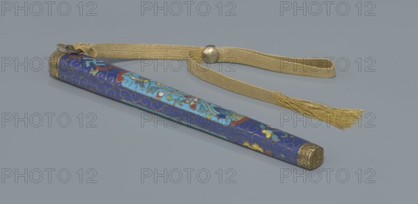 Travel cutlery set in case;, 19th century, China, Qing dynasty. Travel cutlery comprising: a Knife with cloisonné enamel handle and steel blade; b–c Ivory chopsticks decorated with an inlaid black lacquer border ornament at the top; d File decorated similarly to the chopsticks; e Case 28 cm length of cloisonné enamel with bronze fittings engraved at top and bottom, interior lined with wood. The chopsticks and file are held by a band decorated with a matching border; a narrow cotton-silk strap with fringes is attached. Case decorated with floral reserves and plant tendrils in the background. Motifs include plant designs and a swastika. Function: cutlery., A long slender sheath with a decorative blue surface featuring swirling gold outlines and floral-like motifs in lighter blue, red, green, yellow, and white rests on a flat grey background, with ornate metallic gold caps at each end; attached to one end is a woven gold-colored strap with a round metallic bead and a tasseled fringe in go...