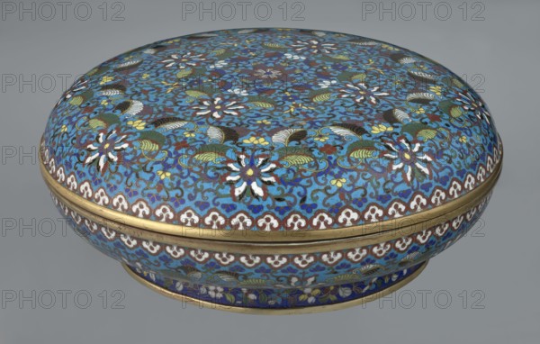 Box with lid, gift, Chinese, Qing dynasty Qianlong period, cloisonné enamel. Large cylindrical-spheroidal box supported on a separate annular foot, with a slightly flattened lid. Covered by a plant tendril encircling the entire surface container and lid, dominated by chrysanthemum flowers and leaves in various stylistic forms; rich multicolored plant composition on a turquoise background. Upper edge of the storage section and lower edge of the cover decorated with a border of ruyi-head motifs in white and red; a blue three-petal pattern completes the decoration. Foot decorated with an ornamental band of colorful flowers and leafy twigs. Interior of the box and inner surface of the foot filled with uniform turquoise enamel. Unglazed edges of the storage section, cover, and foot gilded. 18th century, China. Height 14.5 cm, diameter 34.5 cm., A rounded decorative box with a domed lid covered in an intricate floral and scroll pattern in shades of blue, turquoise, green, white, yellow, brow...