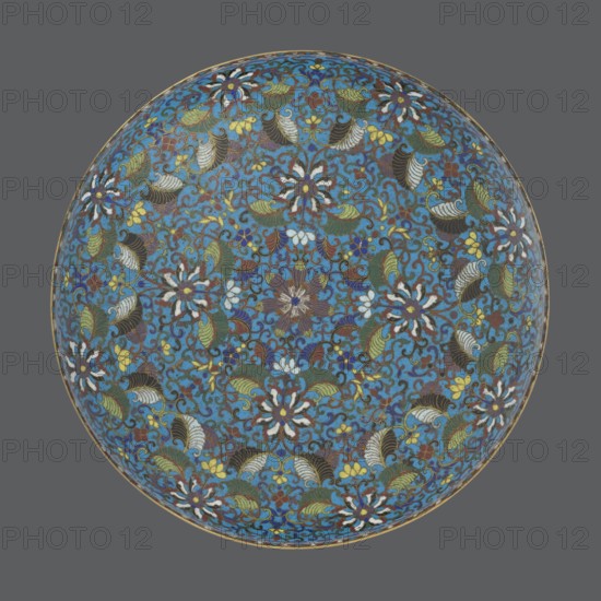 Box with lid, cloisonné enamel, Qing dynasty, Qianlong period, 18th century, China. Large cylindrical-spheroidal box supported on a separate annular foot, with a slightly flattened lid. Surface covered by a plant tendril encircling the entire container and lid, dominated by chrysanthemums and leaves in various stylistic forms against a turquoise background. Upper edge of the storage section and lower edge of the cover decorated with a border of ruyi-head motifs in white and red; a blue three-petal pattern completes the decoration. Foot decorated with a band of colorful flowers and leafy twigs. Interior of the box and inner surface of the foot filled with uniform turquoise enamel. Unglazed edges of the storage section, cover, and foot are gilded. Height 14.5 cm; diameter 34.5 cm., A round decorative object with an intricate floral pattern covering its surface, set against a solid neutral background; the pattern features a dominant blue field with several stylized flowers and leaves in c...