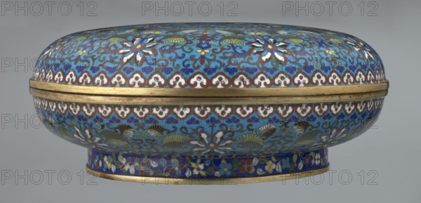 Box with lid, Chinese, Qing Qianlong, cloisonné enamel. Large cylindrical-spheroidal box supported on a separate annular foot, with a slightly flattened lid. Surface covered by a plant tendril encircling the container and lid, dominated by chrysanthemum flowers and leaves in various stylistic forms on a turquoise background. Upper edge of the storage section and lower edge of the cover decorated with a border of ruyi-head motifs in white and red; a blue three-petal pattern completes the decoration. Foot bears an ornamental band of colorful flowers and leafy twigs. Interior of the box and inner surface of the foot filled with uniform turquoise enamel. Unenamelled edges of the storage section, cover, and foot are gilded. 18th century, China. Height 14.5 cm; diameter 34.5 cm., An oval lidded container with a rounded lid and short pedestal base covered in intricate enamel patterns in shades of blue, turquoise, white, green, brown, yellow, and gold; the surface shows repeating floral and sc...