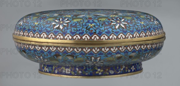 Box with lid, Chinese, Qing dynasty Qianlong period, cloisonné enamel, 18th century. Large cylindrical-spheroidal box supported on a separate annular foot, with a slightly flattened lid. Surface covered by a plant tendril encircling the container and lid, dominated by chrysanthemum flowers and leaves in various stylistic forms, rendered in rich multicolored enamel against a turquoise ground. Upper edge of the storage section and lower edge of the cover decorated with a border of ruyi-head motifs in white and red, with a blue three-petal pattern completing the decoration. Foot bears an ornamental band of colorful flowers and leafy twigs. Interior of the box and inner surface of the foot filled with uniform turquoise enamel; unenamelled edges of the storage section, cover, and foot are gilded. Height 14.5 cm; diameter 34.5 cm., Function: container., A rounded, oval decorative box with a domed lid and a short cylindrical base, covered in intricate floral and scroll patterns. The surface s...