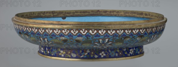 Box with lid, cloisonné enamel, Qing dynasty, Qianlong period, 18th century, China. Large cylindrical, spheroidal box supported on a separate annular foot, with a slightly flattened lid. Surface covered by a plant tendril encircling the entire container and lid, dominated by chrysanthemum flowers and leaves in various stylistic forms against a turquoise background. Upper edge of the storage section and lower edge of the cover decorated with a border of ruyi-heads in white and red; a blue three-petal pattern completes the decoration. Foot has an ornamental band of colorful flowers and leafy twigs. Interior of the box and inner surface of the foot filled with uniform turquoise enamel. Unenamelled edges of the storage section, cover, and foot are gilded. Height 14.5 cm; diameter 34.5 cm., A shallow decorative bowl with a wide rim and low pedestal base, showing intricate floral and scroll patterns in shades of blue, turquoise, white, green, brown, and gold; the interior appears sky blue wi...