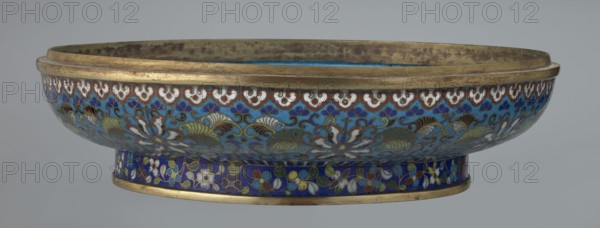 Box with lid, Qing dynasty, Qianlong period, cloisonné enamel, floral ornament featuring chrysanthemum tendrils covering container and lid against a turquoise background. Large cylindrical-spheroidal box on a separate annular foot with slightly flattened lid. Upper edge of the storage section and lower edge of the cover decorated with a ruyi-head border in white and red; a blue three-petal pattern completes the decoration. Foot bears an ornamental band of colorful flowers and leafy twigs. Interior of the box and inner surface of the foot filled with uniform turquoise enamel; unenameled edges storage section, cover, and foot are gilded. 18th century, China, container; height 14.5 cm, diameter 34.5 cm., An oval shallow bowl with a raised foot, decorated in shades of blue, turquoise, white, green, brown, yellow, red, and gold; the outer surface shows a repeating floral and foliate pattern with stylized flowers and leaves in white, green, brown, and yellow against a blue and turquoise back...