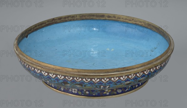 Box with lid. Chinese, Qing dynasty, Qianlong period, 18th century. Cloisonné enamel large cylindrical-spheroidal box supported on a separate annular foot, with a slightly flattened lid. The vessel is covered by a plant tendril encircling the entire surface container and lid, dominated by chrysanthemum flowers and leaves in various stylistic forms, set against a turquoise background. The band at the upper edge of the storage section and the lower edge of the cover are decorated with a ruyi-head border in white and red; a blue three-petal pattern completes the decoration. The foot has an ornamental band of colorful flowers and leafy twigs. The interior of the box and the inner surface of the foot are filled with uniform turquoise enamel. Unground portions edges of the storage section, cover, and foot are gilded. Height 14.5 cm; diameter 34.5 cm., A shallow, round bowl with a bright light blue interior and a metallic gold rim, showing a few small darker spots and subtle surface reflectio...
