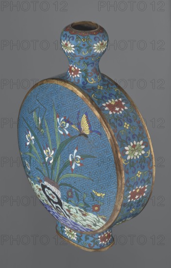Vase - moon bottle pilgrim bottle with a flattened belly, supported by a separate oval high foot, with a short neck resembling a garlic bulb suantoukou type. Decoration on one side: a multicolored composition depicting a chrysanthemum flower growing on a coastal rock, above which a dragonfly and a grasshopper hover. The other side: lilies irises on a rock above the water, with a butterfly and a grasshopper perched above them. Small, colorful flowers growing on the shore are also visible in both representations. Both compositions use the flowers and birds motif known from Chinese scroll painting. A Chinese meander forms the background for both compositions. The foot, side edges, and neck are filled with a lotus tendril pattern. Colors dominated by red, white, green, yellow, and navy blue; elements without enamel are gilded. Visible abrasions of the gilding and firing-related technological defects. Cloisonné enamel. Qing dynasty, 2nd half 18th century. China. Vessel. Height 29.8 cm; widt...