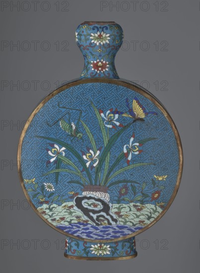 Vase - moon bottle pilgrim bottle with a flattened belly, supported by a separate oval high foot, with a short neck resembling a garlic bulb suantoukou type. Decoration on one side: a multicolored composition depicting a chrysanthemum flower growing on a coastal rock, above which a dragonfly and a grasshopper hover. The other side: lilies irises on a rock above the water, with a butterfly and a grasshopper perched above them. Small, colorful flowers growing on the shore visible in both representations. Motif derived from Chinese scroll painting flowers and birds. A Chinese meander forms the background for both compositions. Foot, side edges, and neck filled with a lotus tendril. Dominant colors red, white, green, yellow, and navy blue. Elements without enamel decoration are gilded; visible abrasions of the gilding and technological firing defects. Cloisonné enamel; moon bottles; Qing dynasty, 2nd half 18th century; Chinese. Vessel. Height 29.8 cm, width 21.1 cm., A round, flat decorati...