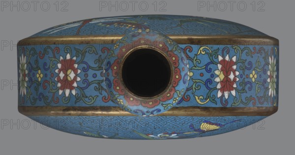 Vase - moon bottle pilgrim bottle with a flattened belly, supported by a separate oval high foot, with a short neck resembling a garlic bulb suantoukou type. Decoration on one side: a multicolored composition depicting a chrysanthemum flower growing on a coastal rock, above which a dragonfly and a grasshopper hover. The other side shows lilies irises on a rock above the water, with a butterfly and a grasshopper perched above them. Small, colorful flowers growing on the shore appear in both scenes. Both decorations use the flowers and birds motif known from Chinese scroll painting; a Chinese meander forms the background for both compositions. The foot, side edges, and neck are filled with a lotus tendril pattern. Cloisonné enamel dominated by red, white, green, yellow, and navy blue; non-enamelled elements are gilded. Visible abrasions of the gilding and firing-related technological defects. Qing dynasty 1644–1911, 2nd half 18th century. Chinese cloisonné vessel. Height 29.8 cm; width 2...