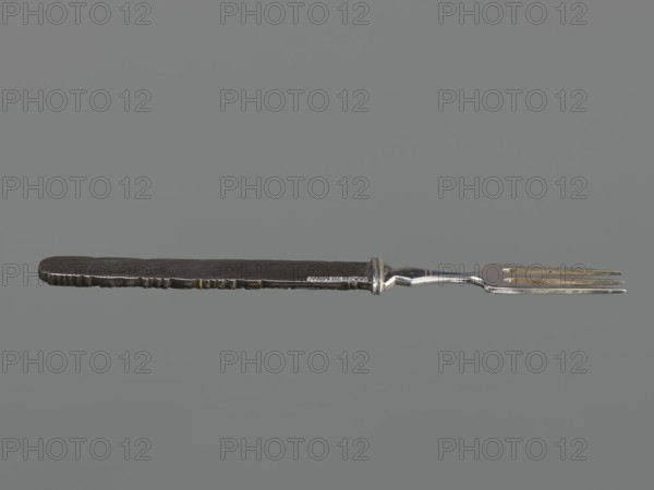 Fork with knife handle kozuka, Japanese, Edo period 1603–1868, 18th century; materials: metal cutlery; subject/iconography: dragons and fish; function: kozuka knife handle for a sword katana; dimensions: height 16.8 cm, width 1.4 cm., A single metal fork with a dark handle and a lighter, silver-toned tined end is shown horizontally against a plain gray background; the handle appears textured with uneven edges and a few small lighter spots, while the central section transitions to a shinier silver connector leading to a three-pronged fork head that shows some darker and slightly yellowed areas.