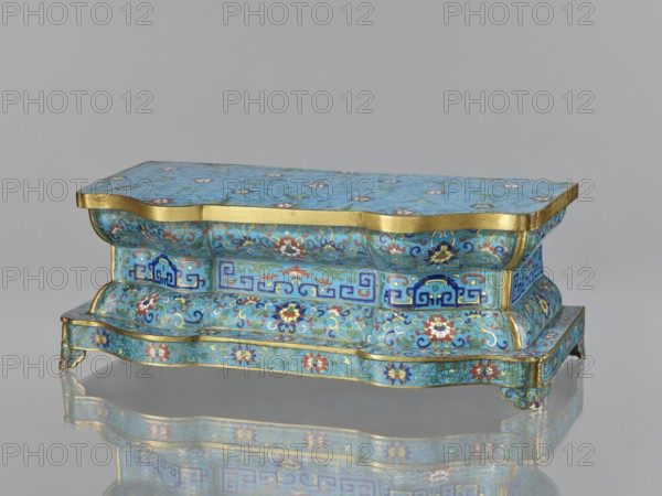 Wall pedestal; creator, cloisonné enamel; decoration: flowers on a turquoise background with a grid of cracks cracked ice, floral tendrils on the profiles, and a central band with geometric motifs; plant and floral and geometric ornament; Qing dynasty 1644–1911, 18th century; China; height 13.5 cm, width 36.6 cm, depth 18.5 cm., A small ornate rectangular box with a wavy, scalloped profile sits on a reflective surface, showing a mirrored underside. The box is covered in a bright turquoise-blue enamel background with intricate floral and foliate patterns in colors including blue, red, white, green, and gold, with gold bands edging the top and base. The sides feature repeating decorative panels with stylized geometric motifs and scrolling vinework, and the top surface displays scattered floral rosettes set against a network of fine crackle lines. The form includes curved, bulging sections and small bracket feet, and the overall appearance is glossy and richly decorated.
