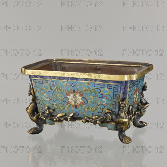 Flower pot incense burner on four legs, 18th century, China, Qing 1644–1911, cloisonné enamel. Decoration: lotus tendril on a turquoise background; legs shaped as flowered trunks plum branches made of gilded bronze. Motifs: lotuses, lotus tendril, plum trees, cracked ice, meander. Function: jardinière / incense burner. Dimensions: height 9.5 cm; width 17.2 cm; depth 12.0 cm., A rectangular decorative container sits on a reflective surface, showing a group of ornate feet with curved shapes in dark metallic and gold tones. The body displays an intricate floral and scrolling pattern in shades of blue, turquoise, green, red, white, and gold, bordered by geometric motifs in blue and gold along the top edge, which is finished with a gold rim. The interior appears darker, with brown and muted metallic hues, and small rivet-like details are visible near the upper inner wall. The overall color palette includes blue, turquoise, green, red, white, gold, brown, and dark metallic tones.
