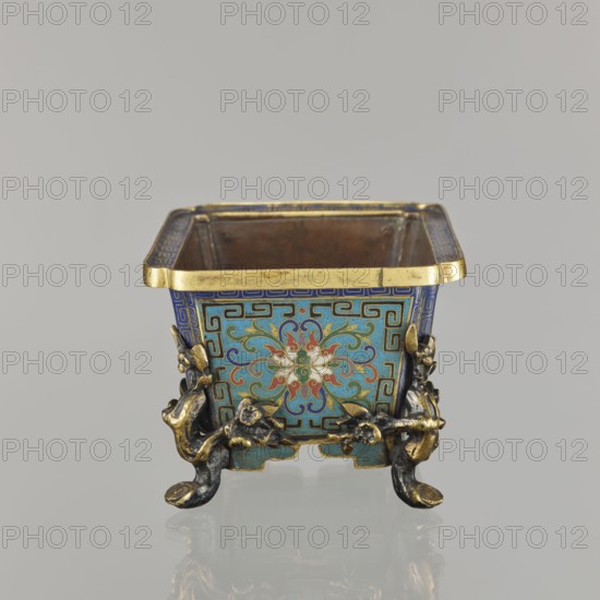 Flower pot incense burner on four legs, cloisonné enamel; decoration: lotus tendril on a turquoise background; legs in the shape of flowered trunks plum branches made of gilded bronze; motifs include lotuses, plum trees, cracked ice, meander; jardinère form; 18th century, Qing 1644–1911, China; vessel; height 9.5 cm, width 17.2 cm, depth 12.0 cm., A small square metal container with an open top sits on a reflective surface, showing a decorative exterior in blue, turquoise, gold, red, white, and green. The upper rim is gold, and each side panel features a central floral motif with swirling petal shapes in red, white, and green set against a turquoise background framed by blue geometric borders. Ornate gilt and dark metal mounts form a group of sculpted elements at the corners and act as feet with curved, claw-like ends. The interior of the container appears plain and metallic with a brownish tone. The background is a uniform light gray.