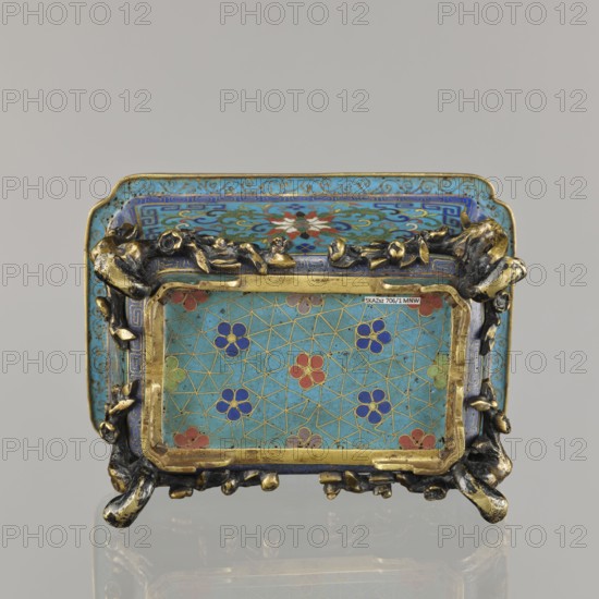 Flower pot incense burner on four legs, Chinese, Qing dynasty 1644–1911, cloisonné enamel, 18th century. Decoration: lotus tendril on a turquoise background; legs in the shape of flowered trunks plum branches made of gilded bronze. Motifs: lotuses, lotus tendril, plum trees, cracked ice, meander. Function: jardinière/incense burner. Dimensions: height 9.5 cm; width 17.2 cm; depth 12.0., A small rectangular decorative object with a turquoise background and a pattern of floral motifs in blue, red, green, and purple set within a gold metal frame; the top surface shows a central multicolored floral rosette surrounded by swirling blue and turquoise ornament. The frame has a textured, organic appearance with dark metallic and gold tones resembling twisted branches or roots at the corners and along the edges. The object sits on a reflective surface that mirrors its colors faintly, and the background behind it is a uniform light gray. The visible colors include turquoise, blue, gold, dark meta...