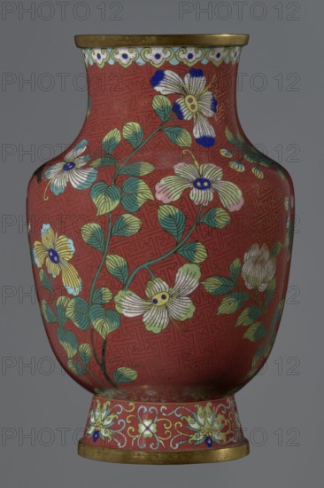 Vase, gift, Chinese art, Chinese culture, Qing 1644–1911, Xianfeng 1851–1861, cloisonné enamel, flowers, floral pattern, meander ornament. The vase is set on a tall, conical foot flaring downwards and terminating in a metal ring, decorated with a lotus tendril. The neck is short and wide, also topped with a metal ring, below which is a border composed of ruyi heads. The belly and neck are decorated with a continuous composition of flowers and plum branches against a red Chinese meander background. 1st half of the 19th century, China. Vessel. Height 25.1 cm; diameter 16.5 cm; weight 1.5 kg., A rounded vase with a narrow neck and flared base, shown against a plain gray background, decorated with a deep red ground covered by a faint geometric pattern and overlaid with floral motifs; there are a few large stylized flowers with white, pale yellow, and pale pink petals tipped with bright blue, and several green leaves with darker green veins arranged on curving stems; a decorative band near ...