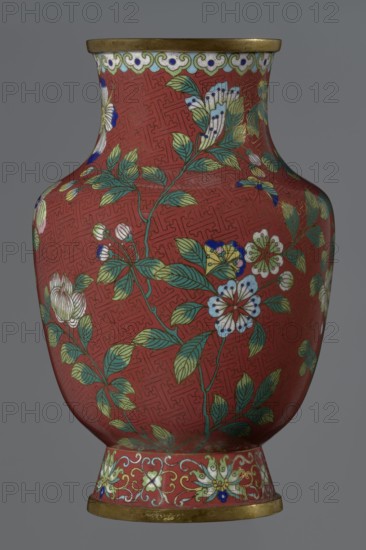 Vase, Chinese; Qing dynasty, Xianfeng period 1851–1861; cloisonné enamel; 1st half of the 19th century. The vase is set on a tall conical foot flaring downwards and terminating in a metal ring, decorated with a lotus tendril. The neck is short and wide, also topped with a metal ring, below which is a border composed of ruyi-heads. The belly and neck are decorated with a continuous composition of flowers and plum branches against a red Chinese meander background. Vessel; height 25.1 cm, diameter 16.5 cm, weight 1.5 kg., A decorative vase with a rounded body and flared neck sits against a gray background; the vase has a rich red ground patterned with a subtle geometric maze-like texture and is decorated with a floral design featuring green leaves and stems, flowers in white, pale yellow, light blue, and touches of dark blue and pink, and small berry-like buds in white and blue; the rim and base edges are metallic gold, and a decorative band near the base includes scrolling floral motifs ...