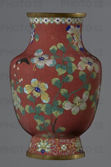 Vase, Chinese, Qing dynasty, Xianfeng period 1851–1861, 1st half of the 19th century. Cloisonné enamel on metal, height 25.1 cm, diameter 16.5 cm, weight 1.5 kg. The vase is set on a tall, conical foot flaring downwards and terminating in a metal ring, decorated with a lotus tendril. The short, wide neck is topped with a metal ring and below it a border composed of ruyi-heads. The belly and neck are decorated all around with a composition of flowers and plum branches against a red Chinese meander background., A rounded vase with a broad neck and flared base, shown against a plain gray background, covered in a red ground patterned with a subtle maze-like design and decorated with floral motifs and foliage. The surface features a group of stylized flowers in white, pale blue, yellow, and cream with blue centers, and stems and leaves in shades of green and teal accented with fine gold outlines. There is a decorative border near the top with pale green and white scalloped elements and smal...