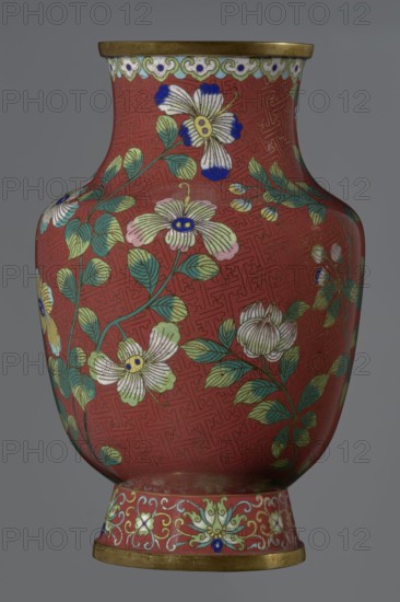 Vase, Chinese, Qing dynasty Xianfeng period, 1851–1861, cloisonné enamel. Tall conical foot flaring downwards terminating in a metal ring, decorated with a lotus tendril. Short wide neck topped with a metal ring, below which is a border of ruyi-heads. Belly and neck decorated in a continuous composition of flowers and plum branches against a red Chinese meander background. 1st half of the 19th century. Vessel; height 25.1 cm, diameter 16.5 cm, weight 1.5 kg., A rounded vase with a narrow rim and flared foot sits against a neutral background, its main surface a deep red decorated with a repeating geometric pattern. Flowing green stems and leaves wind across the red field, punctuated by a group of stylized flowers in pale yellow, white, and light pink with dark blue centers and accents. The rim and foot edges are metallic gold, and the foot band features ornate scrolling motifs in pale green, white, and blue on the same red background. Overall colors visible include red, green, pale yell...